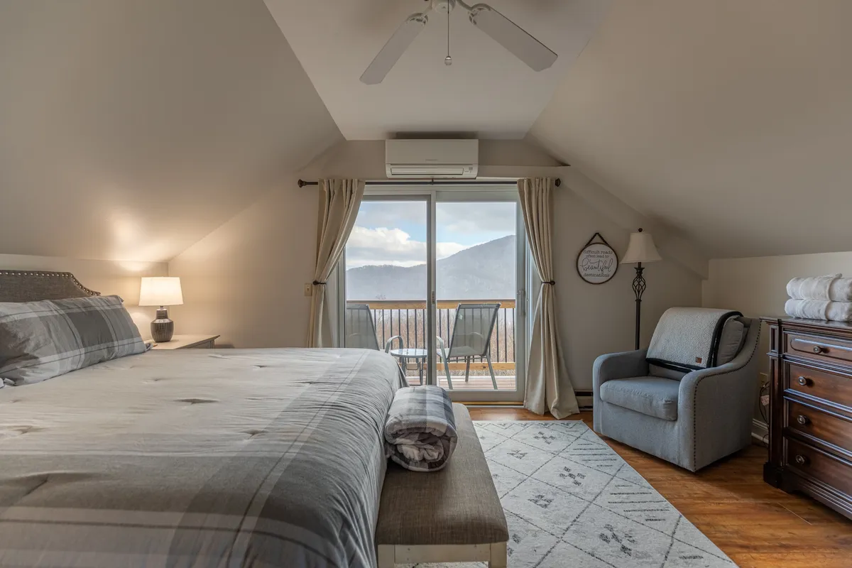 A spacious bedroom featuring a large bed with crisp linens, a comfortable upholstered chair, and a decorative lamp. The room is accented by a panoramic window offering mountain views, creating a serene and private atmosphere ideal for relaxation.