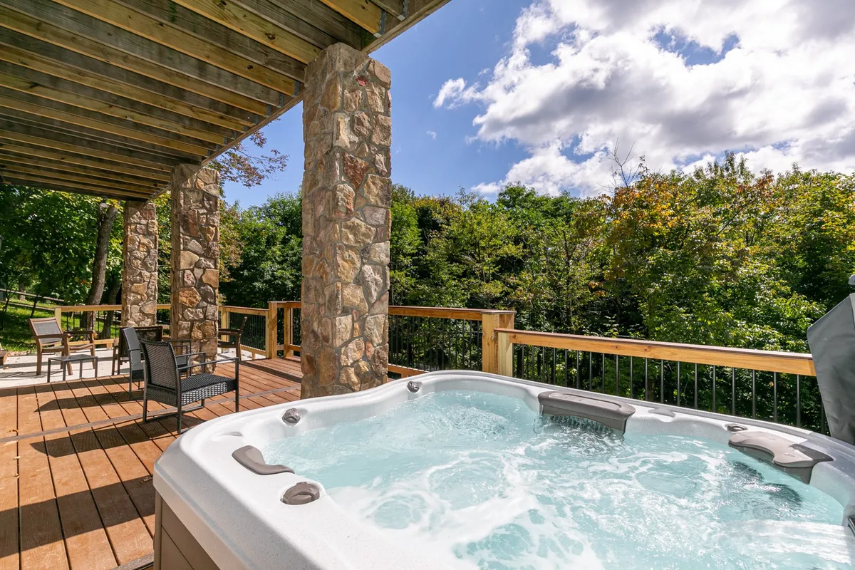 A peaceful outdoor deck surrounded by trees featuring a brand-new hot tub, sturdy stone pillars, and ample wooden decking. The setting is private, inviting, and showcases a relaxing area to soak while immersed in nature.