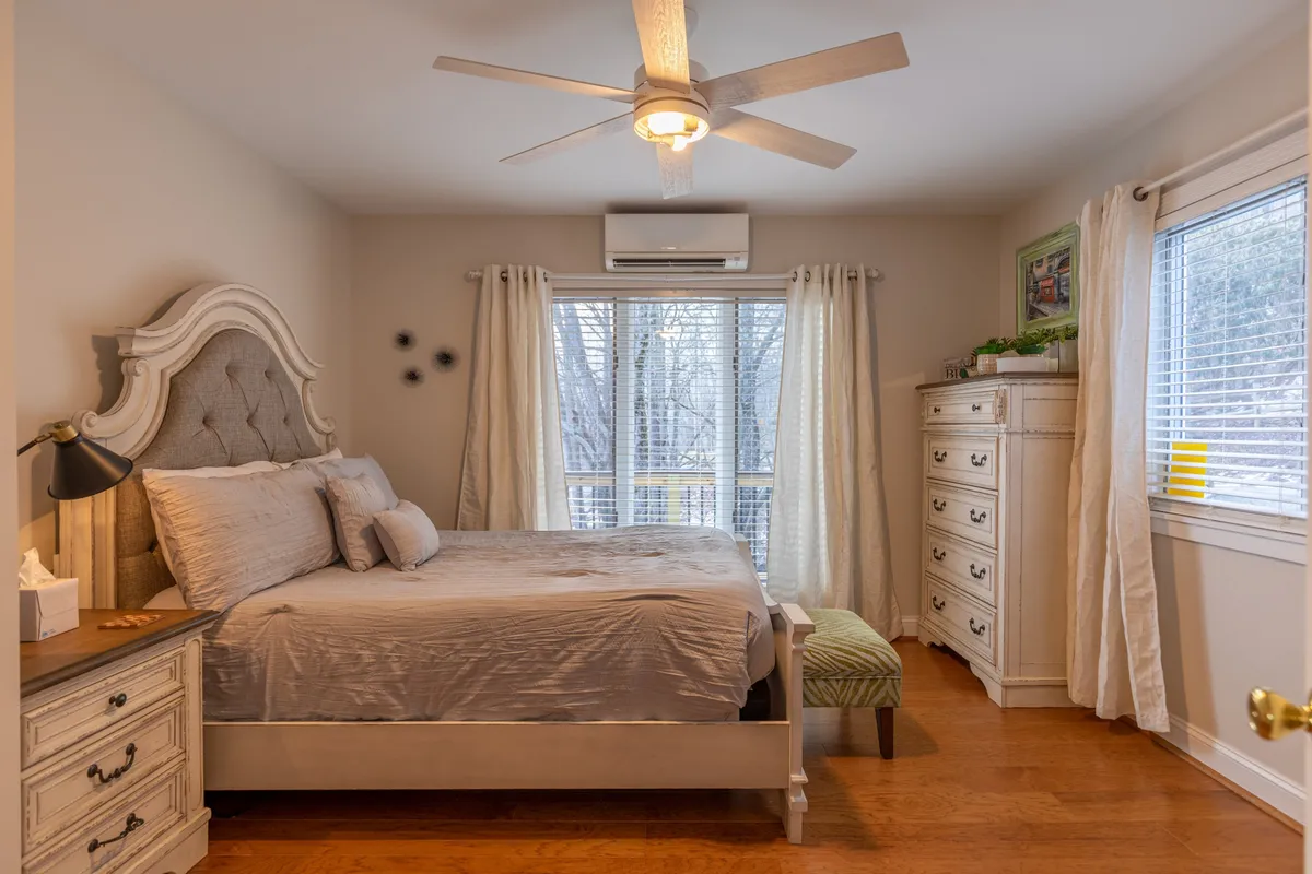 A tranquil bedroom showcasing a neatly made bed, a window letting in natural light with views of surrounding trees, a simple dresser, and a ceiling fan. The understated decor fosters a peaceful and restful environment.