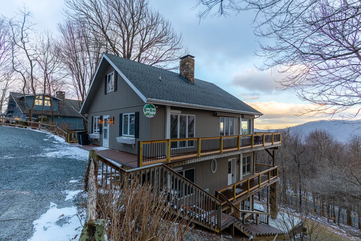 This image features the exterior of Blue Ridge Vista, showcasing the house nestled among lush trees with a spacious deck overlooking expansive mountain views. The inviting deck suggests an ideal spot for relaxing after a hike or enjoying morning coffee amidst nature.