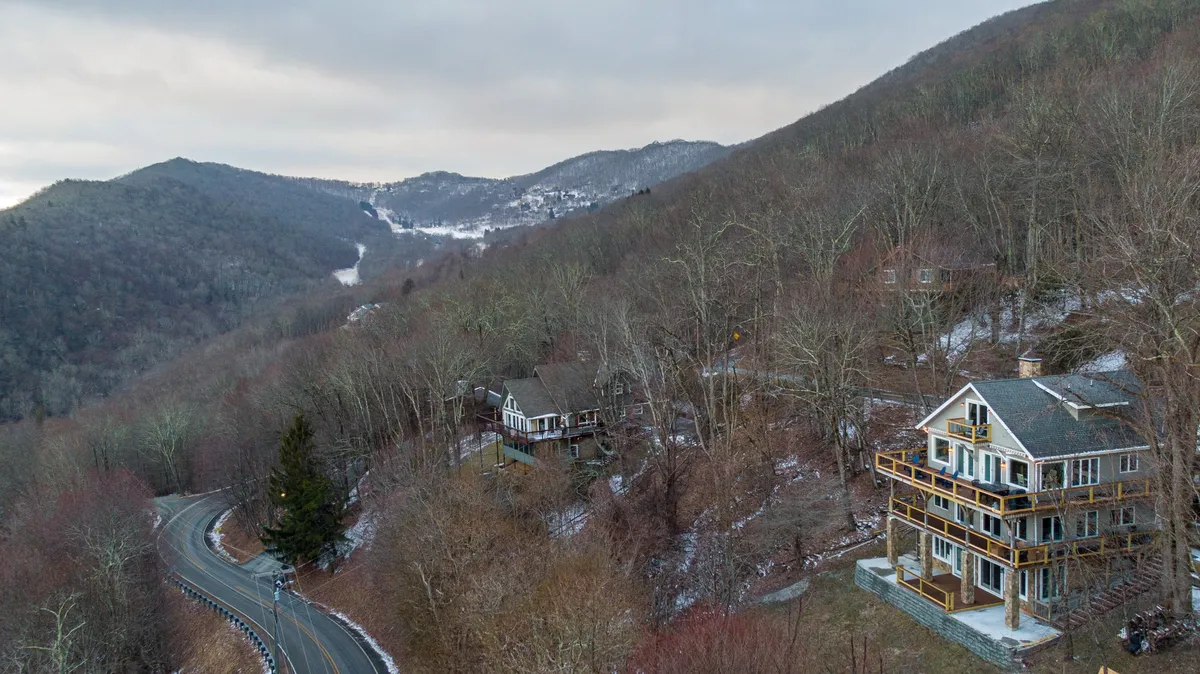 A striking view of a multi-story house nestled in the Blue Ridge mountains, with a curving road leading up to the property. The house features expansive decks that appear to wrap around the structure, and the scene is framed by a panoramic mountainous landscape. The tranquil setting highlights the property's prime location for enjoying outdoor activities and appreciating the natural surroundings.
