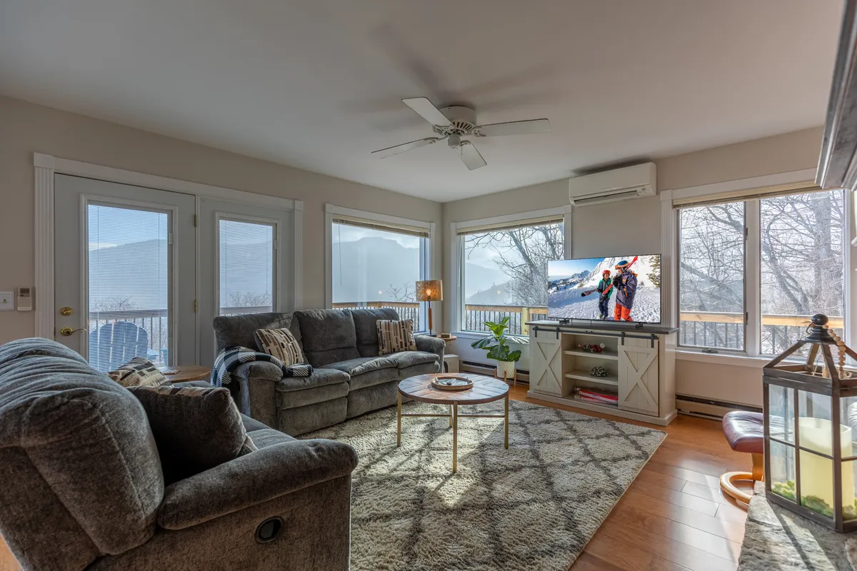 A bright, comfortable interior seating area with large windows framing mountain views. The cozy couch and inviting decor encourage relaxation, and the expansive windows draw the outside scenery in, blurring the line between indoor comfort and natural beauty.
