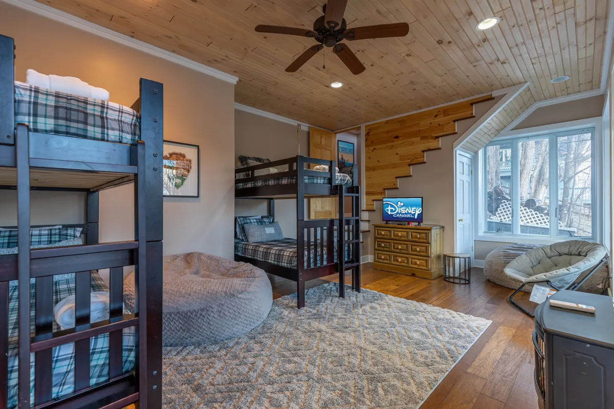 A playful bunk room featuring two sets of bunk beds, a cozy bean bag chair, a television, and a wooden staircase. The large window lets in plenty of natural light, making the space cheerful and perfect for kids or groups.