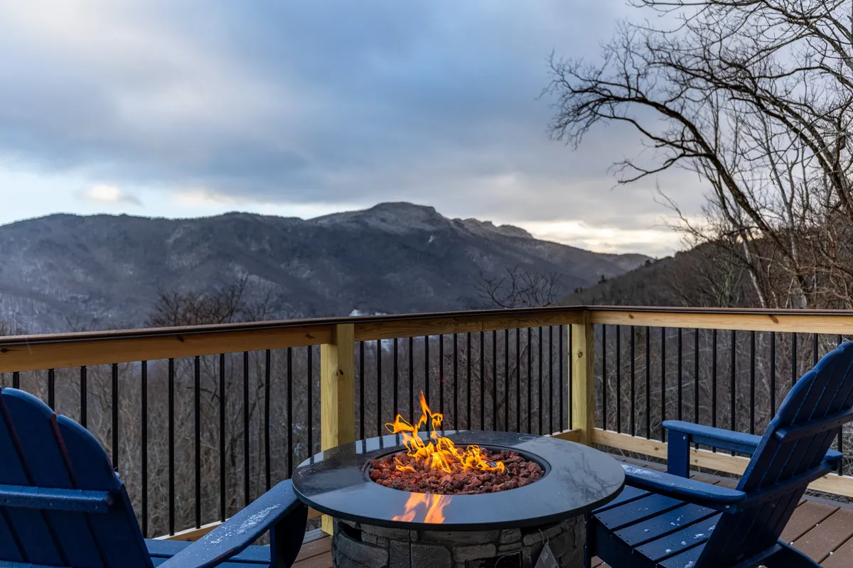 The image captures a cozy fire pit encircled by Adirondack chairs, set on a deck overlooking breathtaking Blue Ridge mountain vistas. The sun is setting beyond the peaks, and firelight glows on the faces of guests gathered, evoking a sense of warmth and togetherness.