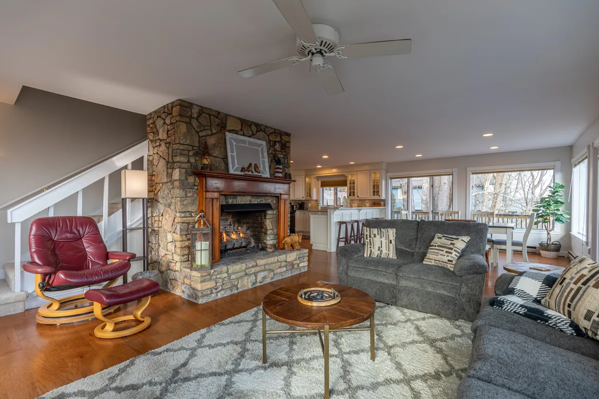 This scene centers on a welcoming living room with comfortable seating, a stone fireplace, and expansive windows that let in abundant light and natural views. The arrangement is ideal for conversations, family nights, or simply relaxing by the fire.