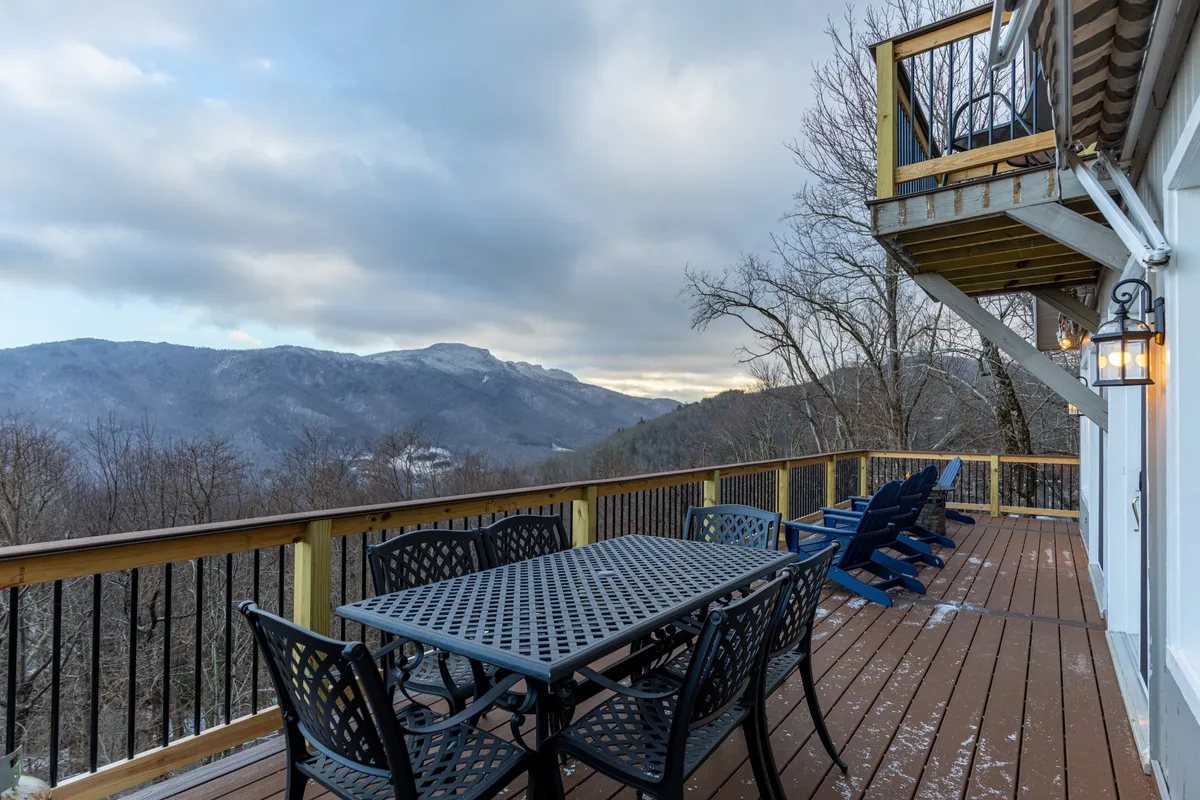 An inviting wrap-around deck equipped with comfortable outdoor furniture overlooks sweeping mountain vistas. The deck’s railing and layout suggest easy access from the main level, perfect for enjoying morning coffee or sunsets by the fire table.